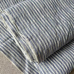Puede incluir: Una pieza de tela doblada con un patrón repetitivo de finas rayas verticales de color azul oscuro sobre un fondo blanco roto. El material presenta un tejido visible y ligeramente texturizado con bordes deshilachados en los pliegues.