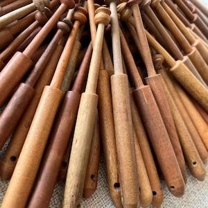 May include: A collection of wooden lace bobbins in various shades of brown. The bobbins have a rounded top and a long, slender body, with a hole at the bottom. The wood grain is visible, and the bobbins appear to be handmade.
