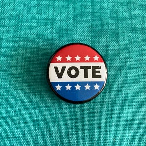 Puede incluir: Un botón rojo, blanco y azul con la palabra "VOTE" en letras negras. El botón tiene un borde negro y está rodeado de una tela turquesa.