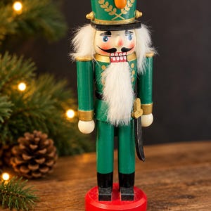 Könnte beinhalten: Eine hölzerne Nussknacker-Figur in einer grünen Uniform mit goldenen Akzenten, einem passenden grünen Hut mit goldenem Emblem und schwarzen Stiefeln. Die Figur hat einen Bart und Haare aus weißem Kunstfell sowie ein schwarzes Schwert als Zubehör.