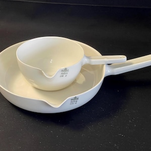 May include: Two white porcelain saucepans with long handles and pouring spouts. One saucepan is nested inside a larger one. The saucepans have a simple, classic design, suitable for cooking and serving. The image is set against a dark background.
