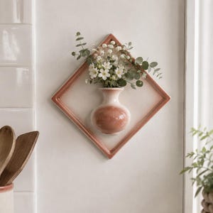 Könnte beinhalten: Eine rautenförmige Keramik-Wandvase mit einem terrakottafarbenen Rand und einem passenden abgerundeten Gefäß in der Mitte. Die Vase enthält einen kleinen Strauß aus weißen Blumen und grünen Eukalyptusblättern und ist flach an einer Wand montiert.