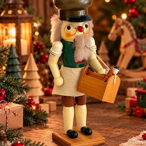Könnte beinhalten: Eine hölzerne Nussknacker-Figur als Bäcker verkleidet, mit dunkelgrüner Jacke, weißer Schürze und brauner Kochmütze. Die Figur hat einen grauen Bart, eine runde Brille und hält einen kleinen hölzernen Werkzeugkasten mit Miniaturwerkzeugen.