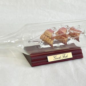 Può includere: Una bottiglia di vetro trasparente contenente una nave a vela in miniatura con tre vele dorate e rosse. La barca poggia su una base di legno scuro con una targhetta dorata con la scritta "Gorch Fock".
