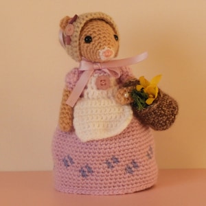 Op de afbeelding: Een gehaakte pop met een roze jurk, een wit schort en een bruin en beige hoedje. De pop houdt een klein bruin mandje vast met gele bloemen.