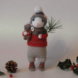May include: A crocheted mouse wearing a red sweater and a white and gray hat, holding a small crocheted bear and a pine branch. The mouse is standing on a white surface with holly and pine cones.