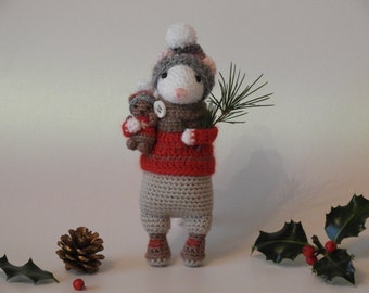 May include: A crocheted mouse wearing a red sweater and a white and gray hat, holding a small crocheted bear and a pine branch. The mouse is standing on a white surface with holly and pine cones.