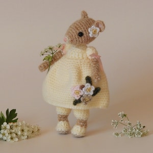 May include: A crocheted stuffed animal in the shape of a mouse wearing a yellow dress with pink flowers and a pink ribbon. The mouse is holding a bouquet of white flowers.