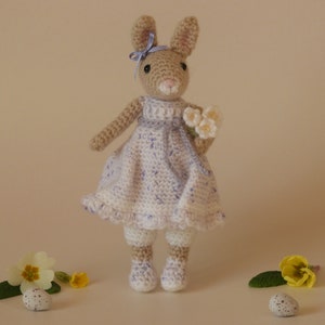 May include: A crocheted bunny doll wearing a white dress with purple accents and holding a bouquet of white flowers. The bunny is standing on a light brown surface with green leaves and yellow flowers.