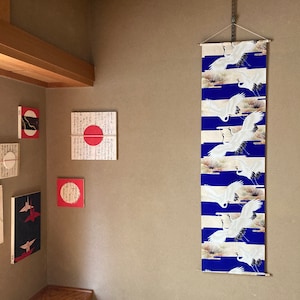 Könnte beinhalten: Ein Wandbehang mit blauem Hintergrund und weißen Kranichen, die in einem Muster fliegen. Die Kraniche sind schwarz umrandet und haben ein goldenes Design. Der Behang ist auf einem Holzstab befestigt und mit einem schwarzen Band aufgehängt.