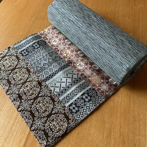 Könnte beinhalten: Ein aufgerollter Stoff mit einem geometrischen Muster in Schwarz, Weiß und Grau. Der Stoff ist über einen braunen Stoff mit einem komplizierteren Muster in Gold, Braun und Weiß gefaltet. Der Stoff liegt auf einer Holzoberfläche.