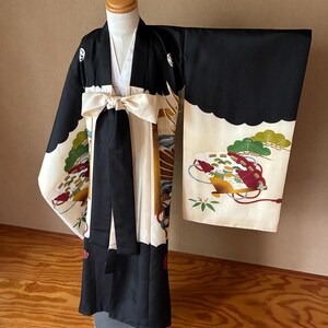 以下が含まれることがあります： 黒と白の着物とクリーム色の帯。着物には、袖と裾にカラフルな花柄と風景画が描かれています。帯は前でリボン結びになっています。マネキンに着物が飾られています。