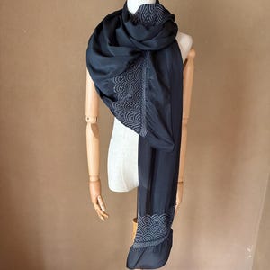 May include: A dark navy blue scarf draped on a wooden mannequin. The scarf has a flowing, silky texture and features a detailed, embroidered wave pattern in a lighter color along the edges. The scarf is long and elegant.