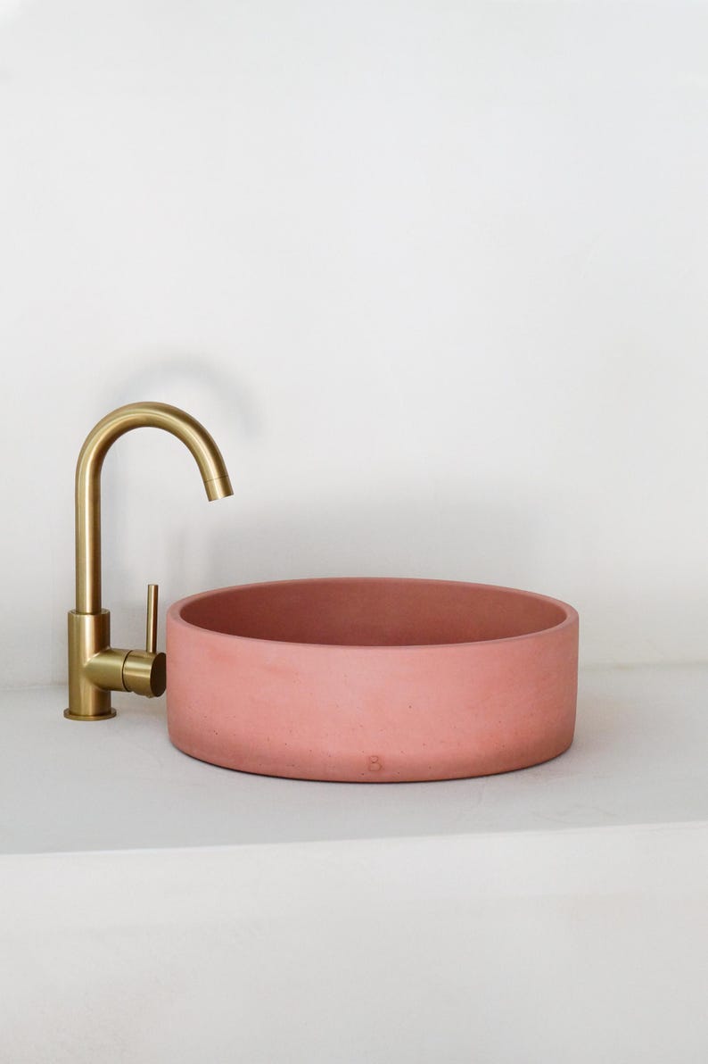 May include: A round, pink concrete vessel sink with a gold faucet. The sink is on a white countertop.