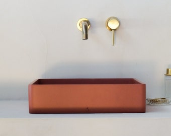 May include: A rectangular, terracotta-colored bathroom sink with rounded edges. The sink is mounted on a white countertop. Two gold-colored faucets are mounted on the wall behind the sink.