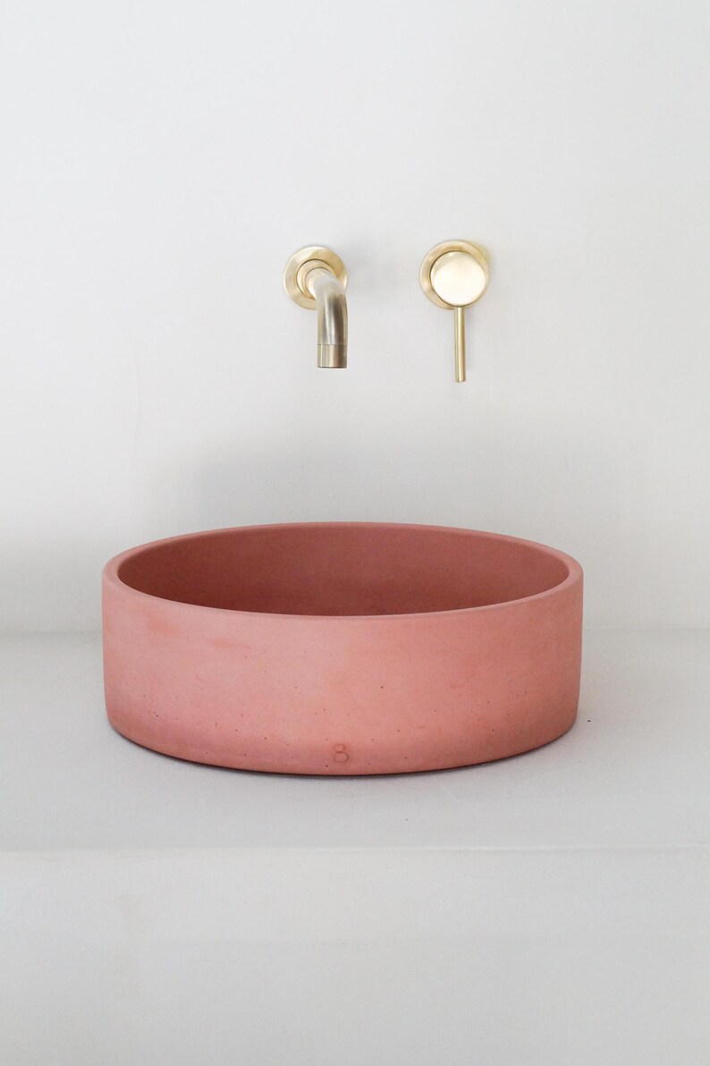 May include: A round, terracotta-colored concrete vessel sink with a brass faucet mounted on the wall above it.