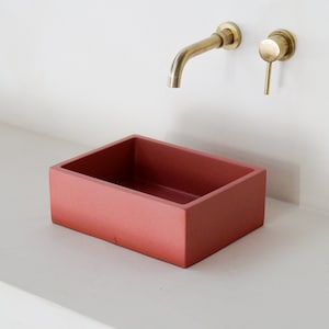 Peut inclure: Un évier rectangulaire en béton de couleur terre cuite. L'évier est posé sur un plan de travail blanc. Un robinet doré est monté sur le mur au-dessus de l'évier.