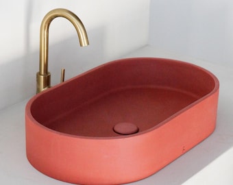 May include: A coral-colored, oval-shaped bathroom sink with a matching drain cover. A gold-colored faucet is mounted on the countertop. The sink has a matte finish and is set against a white wall.