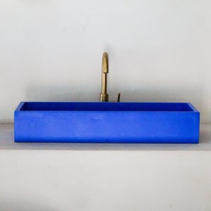 Könnte beinhalten: Ein moderner, rechteckiger, kobaltblauer Waschbecken mit einer Messingarmatur. Das Waschbecken ist leuchtend blau und kontrastiert mit dem metallischen Gold der Armatur. Der Wasserhahn hat einen gebogenen Auslauf und einen einzelnen Griff.