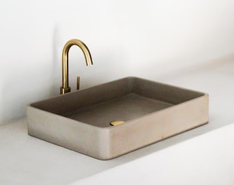 May include: A rectangular concrete bathroom sink in a light beige color. A gold-colored faucet is mounted on the side. The sink has a simple, modern design, suitable for a minimalist bathroom.