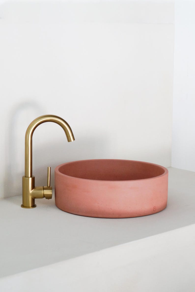May include: A round, terracotta-colored concrete sink with a gold faucet. The sink is on a white countertop.