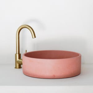May include: A round, pink concrete vessel sink with a gold faucet. The sink is on a white countertop.
