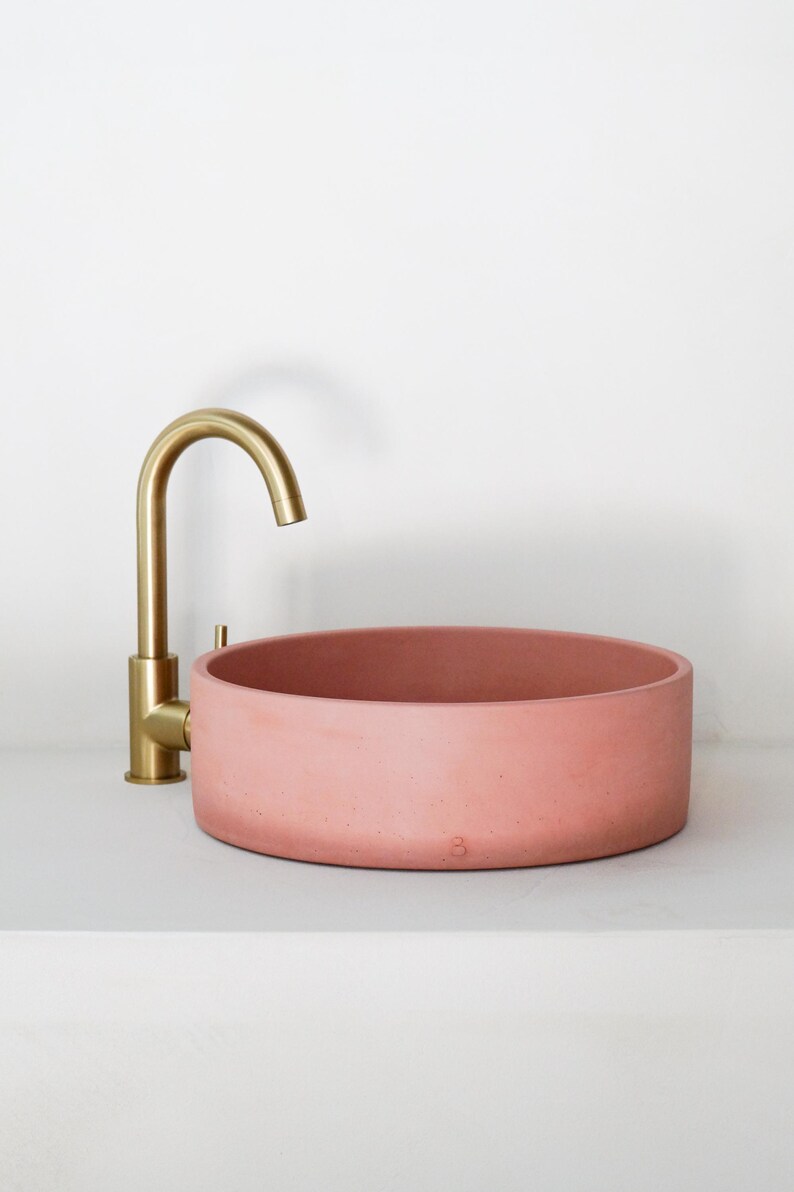 May include: A round, pink concrete vessel sink with a gold faucet. The sink is on a white countertop.