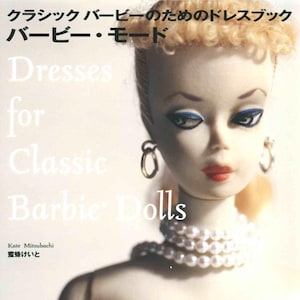 Può includere: Una copertina di libro intitolata Barbie Mode: Dresses for Classic Barbie Dolls con un primo piano di una bambola con capelli biondi, orecchini a cerchio dorati e una collana di perle. Il testo include il nome dell'autrice Kate Mitsubachi e l'editore.