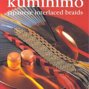 May include: A book cover titled "making kumihimo japanese interlaced braids" with various colorful braided cords and wooden spools. The cover has a red background with a gold-colored plate and a fan.