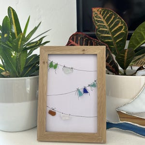 May include: A framed artwork featuring a minimalist design of colorful glass pieces resembling tiny garments hanging on a clothesline. The frame is made of light-colored wood. The artwork is displayed on a white surface, with potted plants in the background.