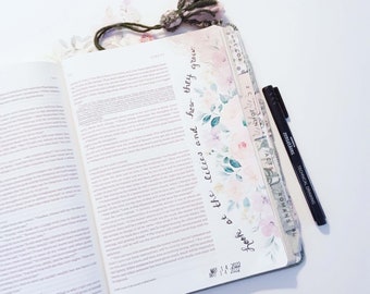 Può includere: Un primo piano di una Bibbia con un segnalibro floreale e una penna nera. Il segnalibro ha il testo "Guarda i gigli e come crescono" scritto su di esso. La Bibbia è aperta al libro di Luca, capitolo 12, versetto 27. La data "14 maggio 2022" è scritta sulla pagina.
