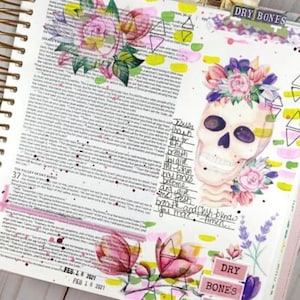 May include: A page from a journal with a watercolor painting of a skull with flowers. The skull is wearing a floral crown and has a pink and white flower on the side. The page is decorated with pink, yellow, and green watercolor paint. The text "DRY BONES" is written on a pink sticker at the top of the page. The text "FEB 18 2021" is written on a pink sticker at the bottom of the page.