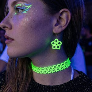 Op de afbeelding: Een vrouw draagt een neon groene choker ketting en bijpassende bloemvormige oorbellen. Haar oogmake-up is neon groen en blauw. Ze draagt een zwarte trui. De achtergrond is een zwak verlichte bar.