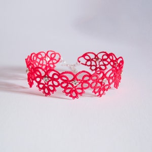 Op de afbeelding: Een rode kanten choker ketting met een bloemenmotief. De ketting is versierd met kleine, sprankelende versieringen. Het delicate kant is gerangschikt in een herhalend patroon, waardoor een mooi accessoire ontstaat.