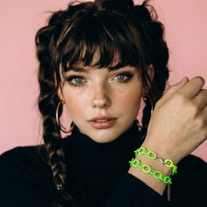 May include: A woman wearing a black turtleneck and a neon green chain bracelet. The bracelet is made of linked circles and has a silver clasp. She has brown hair, blue eyes, and gold hoop earrings. The background is a solid pink.