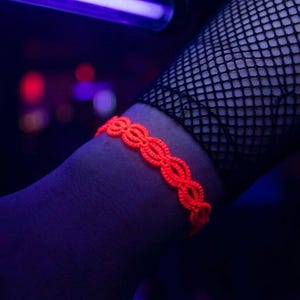 Op de afbeelding: Een neonoranje armband met een open ovaal ontwerp wordt om een pols gedragen. De pols is gedeeltelijk bedekt door een zwarte visnetkous. De achtergrond is donker met hints van rood en paars.