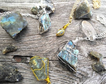 May include: A collection of labradorite necklaces with silver and gold wire wrapping. The raw, iridescent stones are displayed on a rustic wooden surface, showcasing their natural beauty and unique patterns. The necklaces feature delicate chains.
