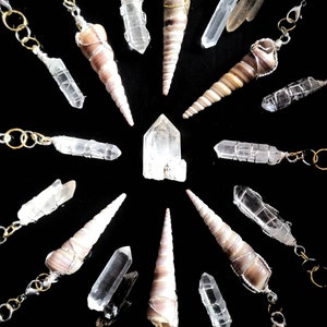 May include: An assortment of crystal pendants and seashell charms on a black background. The pendants feature clear quartz crystals and spiral seashells, some wrapped in silver or gold wire. The charms are connected to silver chains.