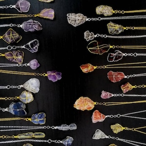 May include: A collection of wire-wrapped crystal necklaces with gold and silver chains. The crystals are various colours and shapes, including clear, purple, red, and green.