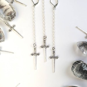 May include: Three silver sword earrings with dangling chains. The swords have intricate details and are attached to silver hoops.