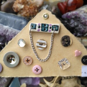 May include: A triangular piece of cardboard with a variety of buttons, beads, and other embellishments. The piece is decorated with a blue and white patterned tile, a silver chain, and the text "T.B.T."