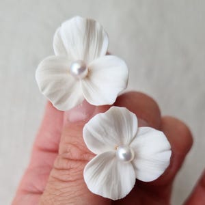 May include: Two white flower-shaped stud earrings, each featuring three delicate, textured petals and a single small, round, lustrous white pearl centred in the middle of the floral design, creating a clean and bright appearance.
