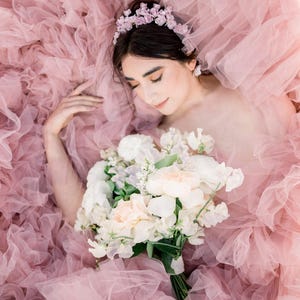 May include: A woman wearing a flower crown lies in a bed of pink tulle fabric, holding a bouquet of white and peach flowers.