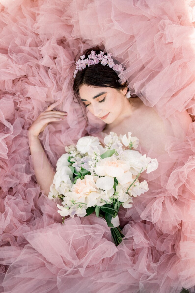 May include: A woman wearing a flower crown lies in a bed of pink tulle fabric, holding a bouquet of white and peach flowers.