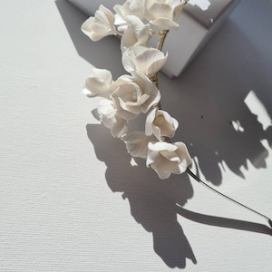 May include: A white floral headband with delicate white flowers and a gold wire base. The headband is on a white surface.