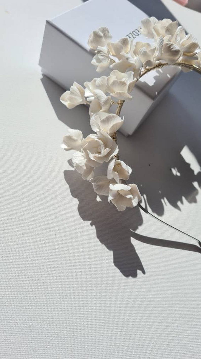 May include: A white floral headband with delicate white flowers and a gold wire base. The headband is on a white surface.