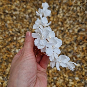 K&ouml;nnte beinhalten: Wei&szlig;er Blumenhaarkamm mit Perlenakzenten. Der Kamm besteht aus wei&szlig;en Stoffblumen und hat einen goldenen Metallkamm.