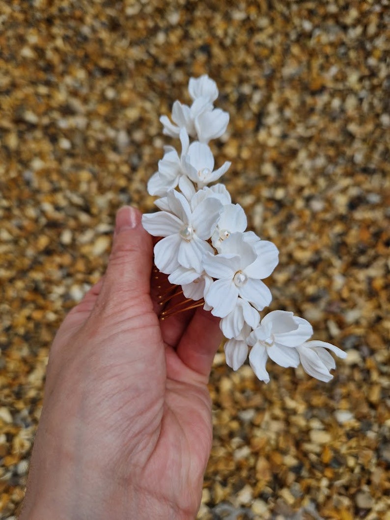 K&ouml;nnte beinhalten: Wei&szlig;er Blumenhaarkamm mit Perlenakzenten. Der Kamm besteht aus wei&szlig;en Stoffblumen und hat einen goldenen Metallkamm.