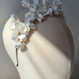 May include: A white floral headband with delicate white flowers arranged in a cascading design. The headband is made of a thin wire and is designed to be worn on the head.