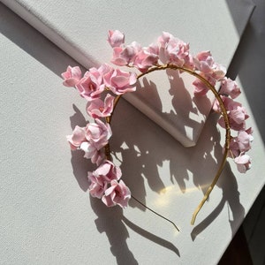 May include: A pink flower crown headband with a gold wire base. The headband is made of delicate pink flowers and is perfect for a special occasion.